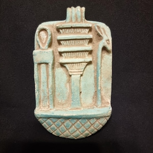 Può includere: Un ciondolo rettangolare turchese con simboli dell'antico Egitto. Il ciondolo presenta un design a colonna centrale, un simbolo Ankh e una figura animale stilizzata. Il fondo ha un motivo a diamante.