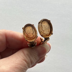 May include: A pair of vintage clip-on earrings. Each earring features an oval, amber-colored cameo with a profile of a woman. The cameos are set in gold-toned metal frames. The earrings are held by a hand against a white background.
