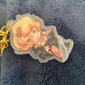 K&ouml;nnte beinhalten: Ein klarer Acryl-Schl&uuml;sselanh&auml;nger mit einer Cartoon-Illustration eines Charakters mit blonden Haaren, der ein schwarzes Outfit mit roten Akzenten tr&auml;gt. Der Schl&uuml;sselanh&auml;nger hat einen goldfarbenen Metallverschluss.