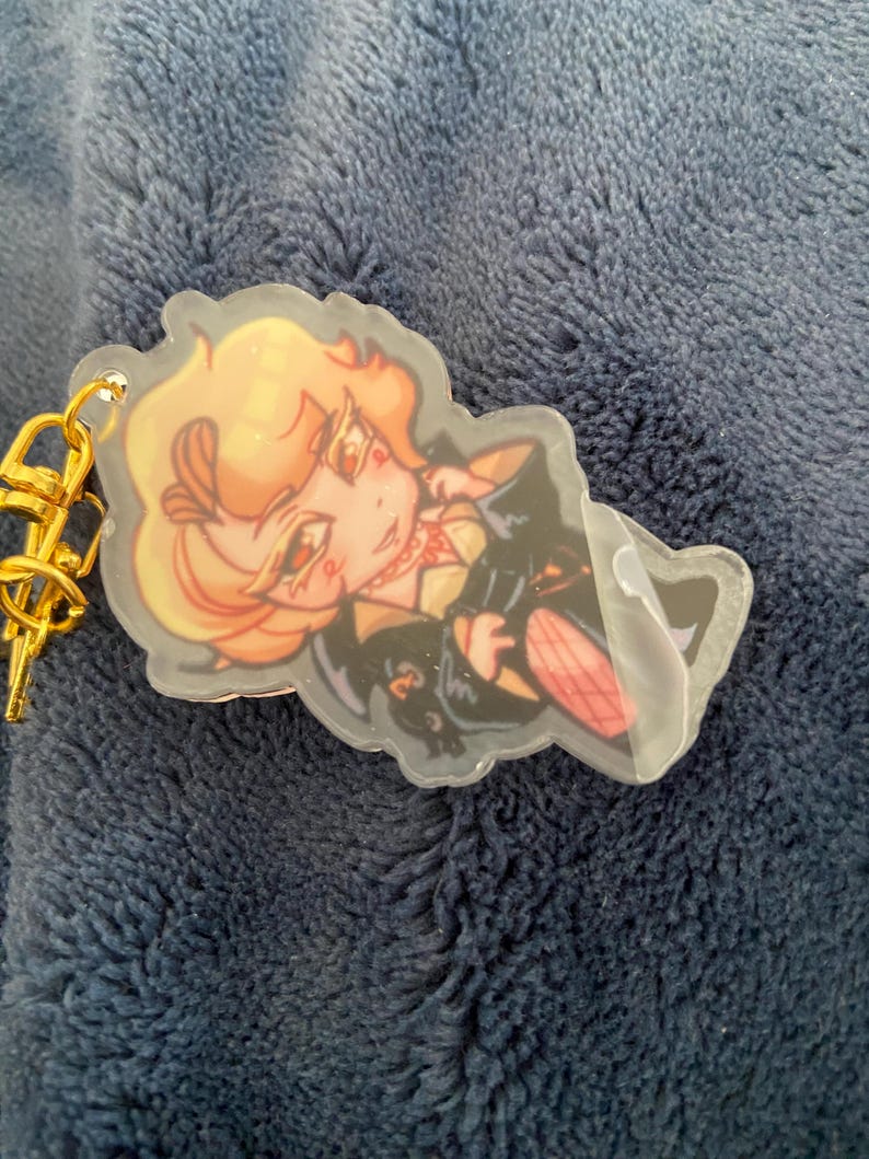 K&ouml;nnte beinhalten: Ein klarer Acryl-Schl&uuml;sselanh&auml;nger mit einer Cartoon-Illustration eines Charakters mit blonden Haaren, der ein schwarzes Outfit mit roten Akzenten tr&auml;gt. Der Schl&uuml;sselanh&auml;nger hat einen goldfarbenen Metallverschluss.