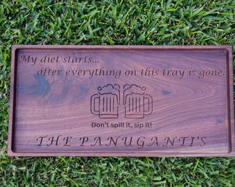 Peut inclure: Un plateau de service rectangulaire en bois avec une finition marron foncé, gravé avec le texte "My diet starts... after everything on this tray is gone." et "Don't spill it, sip it !" avec deux chopes de bière et le nom "THE PANUGANTI'S".