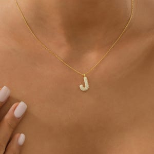 Op de afbeelding: Een goudkleurige ketting met een hanger in de vorm van de letter "J". De hanger is bezet met kleine, sprankelende steentjes. De ketting wordt getoond op de hals van een persoon.