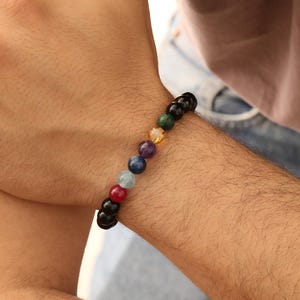 May include: A beaded bracelet featuring a series of colorful gemstones, including red, blue, green, and yellow, strung together with black beads. The bracelet is worn on a wrist, showcasing the vibrant colors and textures of the stones.