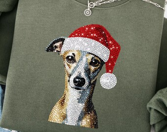 May include: Olive green sweatshirt featuring a rhinestone-studded dog wearing a Santa hat. The dog's portrait is the focal point, with a silver necklace draped across the top. The sweatshirt is folded, with the sleeves and a pair of jeans visible.