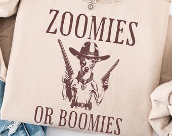 Puede incluir: Sudadera color arena con el texto "ZOOMIES OR BOOMIES" en marrón. El gráfico presenta un perro con sombrero de vaquero sosteniendo dos pistolas. Se ve un collar plateado con un colgante de sol.