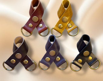 May include: Five leather keychains in various colors: burgundy, mustard yellow, dark brown, purple, and black. Each keychain features a loop design with a metal ring and is secured with two brass-colored rivets.