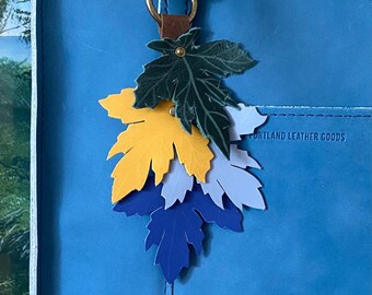 May include: A leather leaf keychain with a brass ring attached to a blue leather bag. The keychain features layered leaves in shades of green, yellow, light blue, and dark blue. The bag is a solid teal color with the words "PORTLAND LEATHER GOODS" printed on it.
