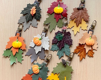 May include: A collection of handmade leather leaf keychains in various autumnal colors. Each keychain features layered leaf shapes, a small pumpkin charm, and a metal clasp. The keychains are in shades of orange, green, brown, and gray.