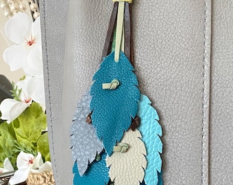 May include: A leather leaf charm in shades of blue, teal, and green, attached to a gray leather bag. The charm has a gold-tone clasp and a yellow accent. The leaves are detailed with stitching.