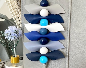 May include: A decorative keychain with a series of layered, bow-shaped leather pieces in shades of gray and blue, interspersed with white and blue beads. The keychain is attached to a gray leather bag with a gold-colored clasp.
