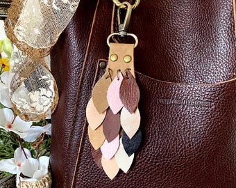 May include: A brown leather bag with a leather tassel keychain. The keychain features a series of leather leaves in various shades of brown, pink, and beige.
