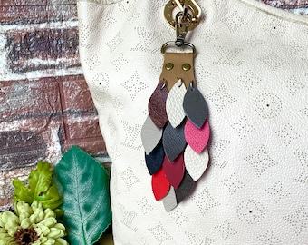 May include: A white leather handbag with a gold chain and a multi-colored leather tassel keychain. The keychain features a series of leather leaves in shades of red, pink, brown, and gray.