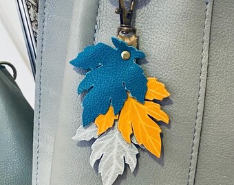 May include: A teal, orange, and white leather leaf keychain charm attached to a silver key ring. The charm is hanging from a gray leather bag.