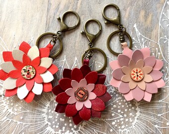May include: Three leather flower keychains in shades of red, pink, and beige. Each flower has multiple layers of petals and a decorative button center. The keychains have bronze-colored clasps and key rings.