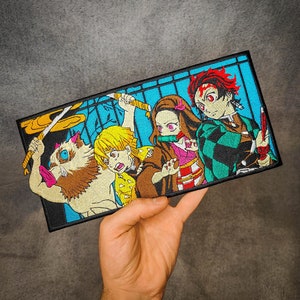 Puede incluir: Parche bordado con cuatro personajes del anime Demon Slayer. El parche es negro con fondo azul y presenta una criatura de color canela similar a un lobo, un personaje de cabello amarillo con una espada, un personaje de cabello rosado con una espada y un personaje de cabello rojo con una espada.