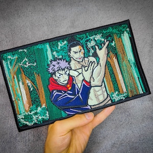 May include: Embroidered patch depicting two men in a woodland scene. One man has purple hair and is wearing a blue and red jacket. The other man has brown hair and is wearing a white shirt. The background is a green forest with trees.