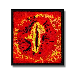 Puede incluir: Parche bordado con un diseño de ojo estilizado. El ojo es principalmente rojo con acentos amarillos y negros, sobre un borde cuadrado negro. El diseño tiene una apariencia texturizada y cosida.