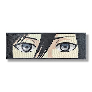 Könnte beinhalten: Gestickter Aufnäher mit einer Nahaufnahme eines Paares blauer Augen mit schwarzen Wimpern und Augenbrauen. Die Augen sind vor einem hellbeigen Hintergrund mit schwarzen Haaren, die das Gesicht umrahmen, platziert.