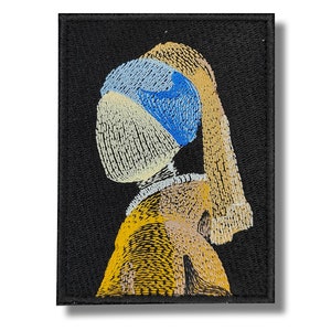 Puede incluir: Parche bordado que representa una pintura famosa, mostrando a una mujer con un pañuelo azul y un pendiente de perla. El fondo es negro, y el rostro de la mujer está parcialmente oculto por el pañuelo.