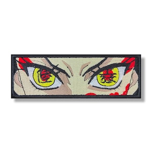 Puede incluir: Parche bordado con un primer plano de un par de ojos amarillos con marcas rojas. Los ojos están rodeados por un contorno negro y un fondo beige.