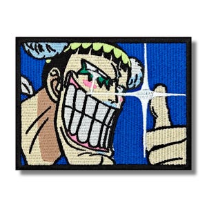 May include: Embroidered patch depicting a cartoon character with a large grin and shining eyes. The character has pink cheeks, green hair, and a blue background. The patch is framed by a black border.