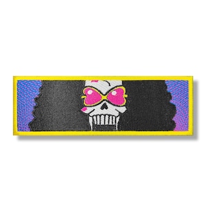 Puede incluir: Una calavera blanca y negra con gafas de sol rosas y un fondo morado y azul. La calavera lleva una peluca afro negra. La imagen es un parche con un borde amarillo.
