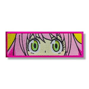 Może przedstawiać: Naszywka haftowana przedstawiająca zbliżenie oczu postaci z kreskówki anime. Oczy są duże i zielone z białymi źrenicami. Postać ma różowe włosy i jasnożółte tło.