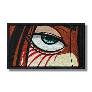 May include: Embroidered patch featuring a detailed eye with a teal iris and a light-colored sclera. The design includes brown hair and red lines, all set against a black border. The patch is rectangular.