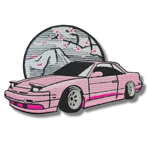 May include: Pink embroidered patch of a Japanese car with a mountain and cherry blossoms in the background.