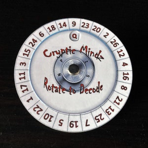 May include: A white and silver cipher wheel with the text "Cryptic Mindz" and "Rotate to Decode" in red. The wheel has numbers around the edge and a rotating dial in the center. A small "Q" is visible.