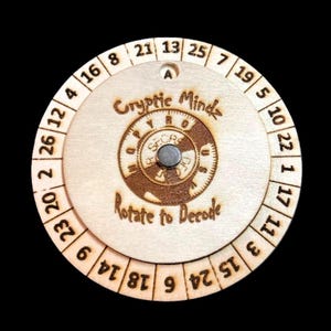 May include: A wooden cipher wheel with engraved numbers and text. The center circle reads "Cryptic Mindz" and "Rotate to Decode". The wheel has a rotating inner disc with a magnetic center. Numbers 1-26 are around the outer edge.