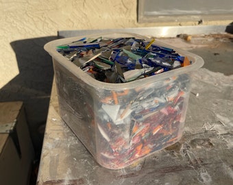 May include: A clear plastic container filled with colorful glass shards. The glass pieces are various shapes and sizes, with colors including blue, green, orange, and silver. The container sits on a metal surface.
