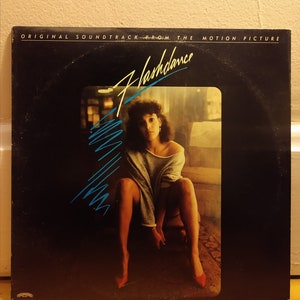 May include: A black vinyl record album cover with the title "Flashdance" in blue and white lettering. The cover features a photo of a woman wearing a grey sweater sitting on a wooden floor.