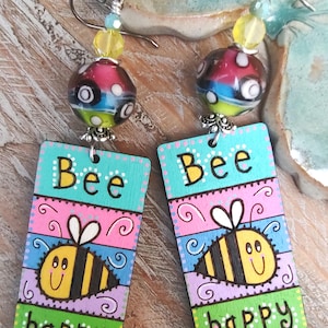 May include: Hand-painted wooden earrings with a bee theme. Each earring features a rectangular pendant with the word "Bee" and "happy" painted on it, along with a cartoon bee. The earrings have colorful accents and beaded tops.