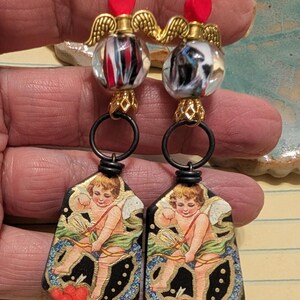 May include: A pair of dangle earrings featuring a cherub design. The earrings have gold-colored angel wings, red heart accents, and black hoops. The main design element is a vintage-style illustration of a cherub with a bow and arrow.