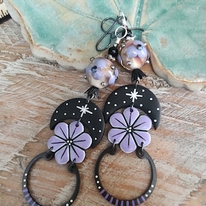 May include: A pair of dangle earrings with a crescent moon and flower design. The earrings are made of black and white clay with purple flowers and white stars. The earrings are attached to silver hoops with a purple and white striped design.