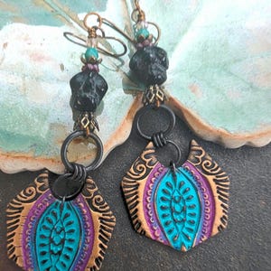 May include: A pair of handcrafted earrings with a bohemian design. The earrings feature black skull-shaped beads, turquoise and purple accents, and ornate, gold-colored metalwork. The focal point is a turquoise teardrop shape with a gold and purple border.