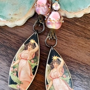 May include: A pair of dangle earrings featuring a pink glass bead and a teardrop-shaped pendant with an image of an angel holding a star. The pendant is black with gold accents.