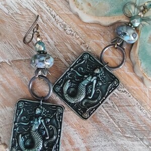 May include: A pair of mermaid themed earrings with a square charm featuring a mermaid with a blue tail and a green body. The charms are attached to a silver hoop and a silver chain with a blue and green glass bead.