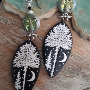 May include: A pair of black and white dangle earrings with a moon and pine tree design. The earrings feature a green iridescent glass bead with a face design.