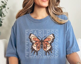 May include: A blue jean colored t-shirt with a butterfly graphic. The butterfly is brown, orange, and cream colored. The text on the shirt reads "I don't know how it gets better than the Fearless" and "You take my hand and drag me head first."