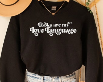 May include: Black sweatshirt with the text "Books are my love language" in white retro font. The sweatshirt is worn with denim shorts and a straw bag.