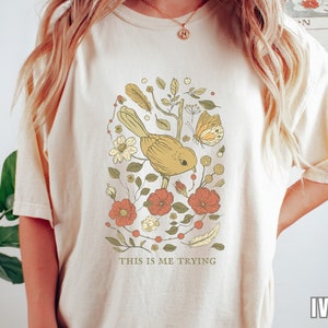 以下が含まれることがあります： クリーム色のTシャツに、黄色の鳥、蝶、赤い花が描かれた花柄のデザインが施されています。デザインの下には「This is me trying」という文字が印刷されています。