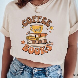 May include: A beige t-shirt with the text "Coffee and Books" in brown and yellow lettering. A yellow coffee cup with a brown handle sits on top of a stack of books. The books are brown with yellow and orange accents. Stars are scattered around the text and the coffee cup.