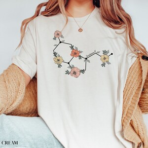 May include: A white t-shirt with a floral graphic of the chemical structure of serotonin. The graphic is in black, pink, and yellow.