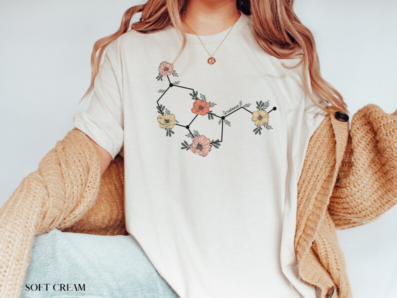 May include: A white t-shirt with a floral graphic of the chemical structure of serotonin. The graphic is in black, pink, and yellow.