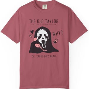 May include: Dusty rose t-shirt featuring a graphic of a ghost-like figure holding a pink telephone. Text includes "THE OLD TAYLOR" and "WHY?" with the phrase "OH, 'CAUSE SHE'S DEAD".