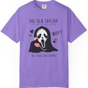 May include: A lilac-colored t-shirt featuring a graphic of the Ghostface mask from the Scream movies holding a pink telephone. Text includes "THE OLD TAYLOR", "WHY?", and "OH, 'CAUSE SHE'S DEAD". The design incorporates hearts.