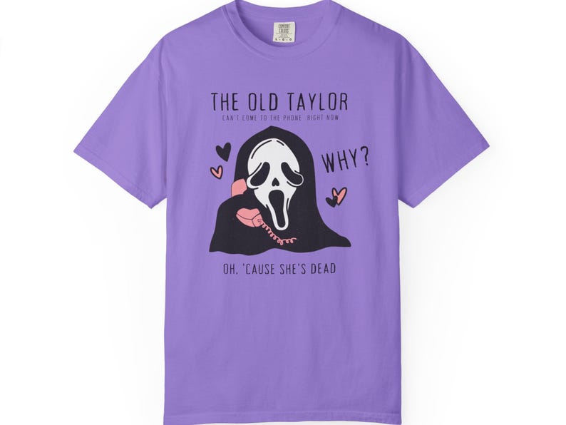 May include: A lilac-colored t-shirt featuring a graphic of the Ghostface mask from the Scream movies holding a pink telephone. Text includes "THE OLD TAYLOR", "WHY?", and "OH, 'CAUSE SHE'S DEAD". The design incorporates hearts.