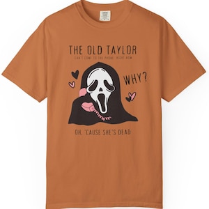 May include: A rust-colored t-shirt featuring a graphic of a ghost-faced figure holding a pink telephone. Text includes "THE OLD TAYLOR" and "WHY?" with hearts. The shirt's design is a playful take on a horror theme.
